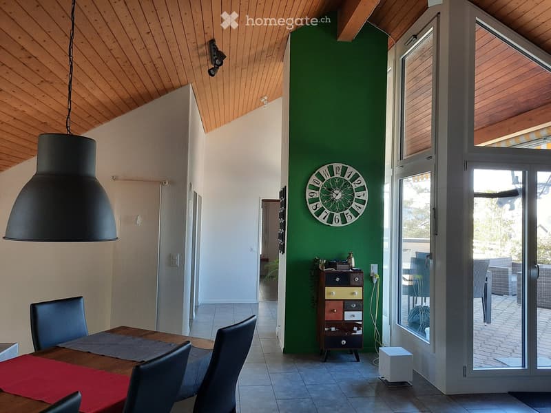 Bright dining area with wooden table, chairs, colorful cabinet, and a large clock on green wall. Large windows and sliding doors lead to outdoor space.