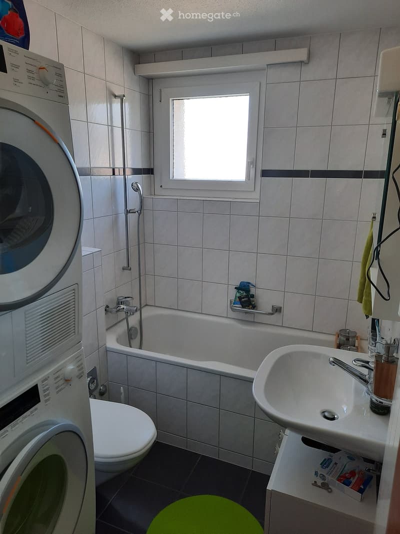 Bathroom with a toilet, bathtub, sink, and washing machine. Features a window above the bathtub and a green stool in front of the sink.