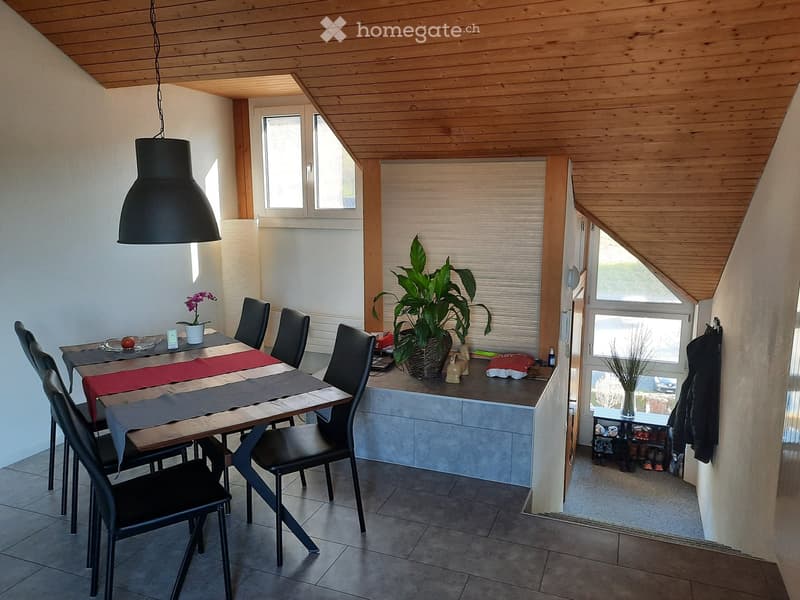 Dining area with a wooden dining table, six black chairs, a large pendant light, and a potted plant on the counter. The room features a slanted wooden ceiling and large windows with blinds.