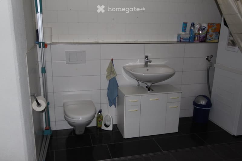 Bathroom with white tiles, toilet, sink, cabinet, and a towel hanging on the wall.