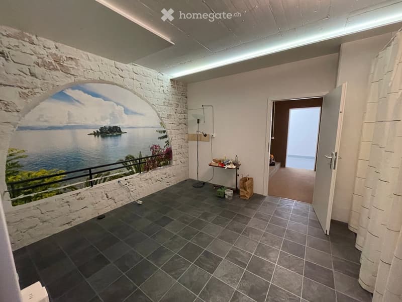 A modern, well-lit room with a large mural of a scenic landscape, a small table with a chair, and a floor lamp. The room features dark tiled flooring and an open doorway leading to another space.