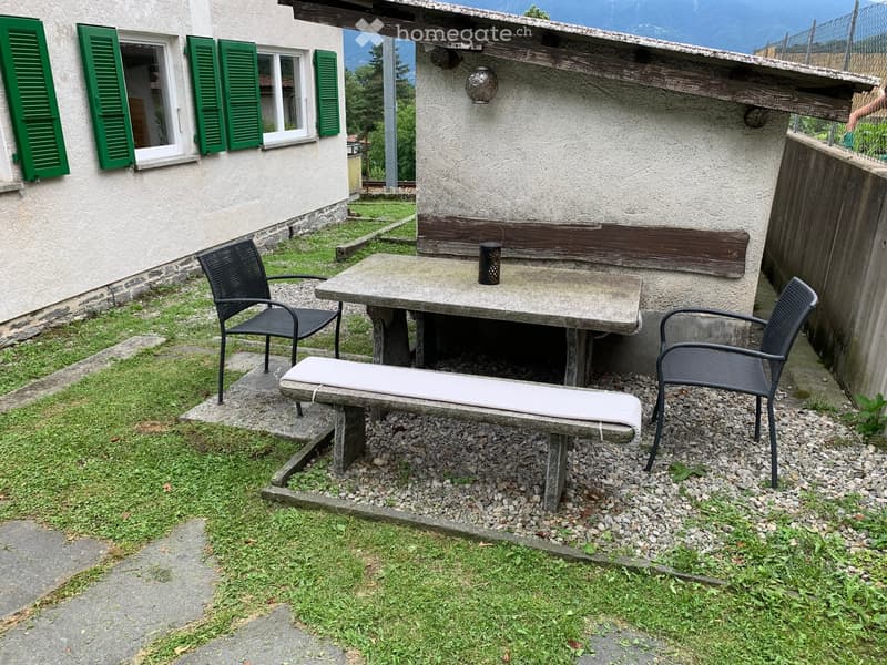 Outdoor seating area with a stone table, two black metal chairs, and a stone bench with a cushion, situated near a house with green shutters and a wooden balcony.