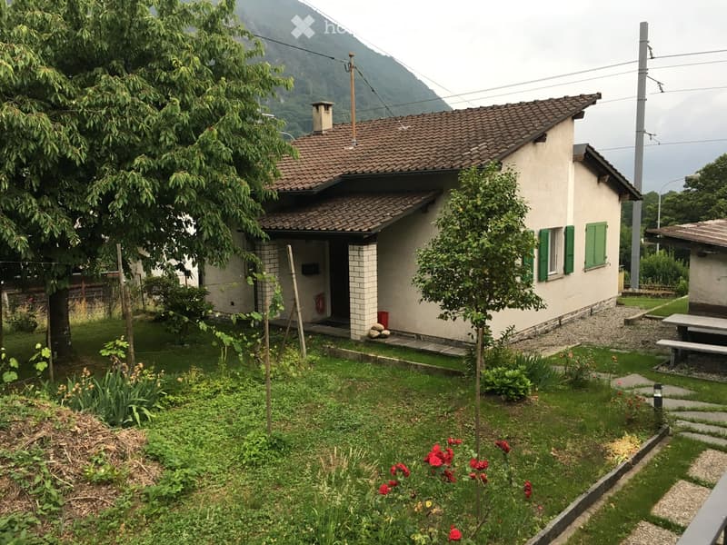 A small, single-story house with a tiled roof and green shutters, surrounded by a well-maintained garden with grass, flowers, and trees. The house has a small porch and is situated in a rural area with mountains in the background.