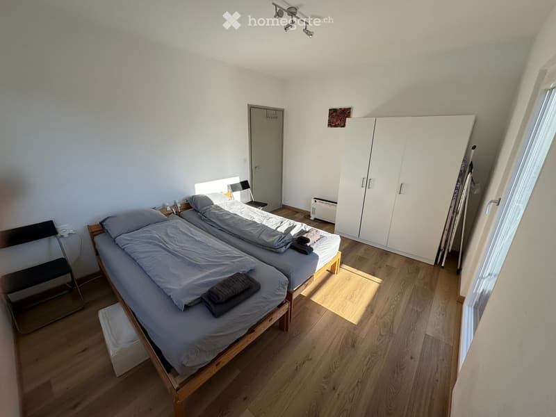 Cozy bedroom with two single beds, a wooden wardrobe, a chair, and a radiator. The room features light wooden flooring and white walls.