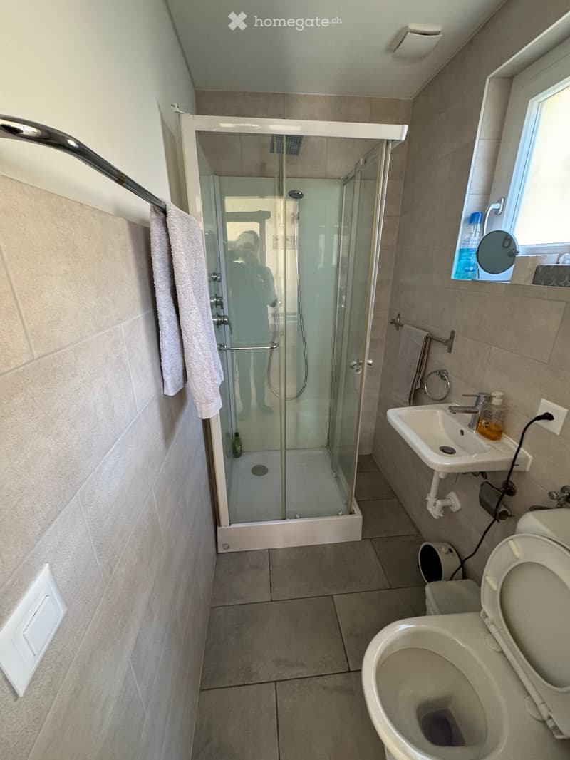 Modern bathroom with a glass-enclosed shower, toilet, sink, and towel. Light-colored tiles and a window provide natural light.