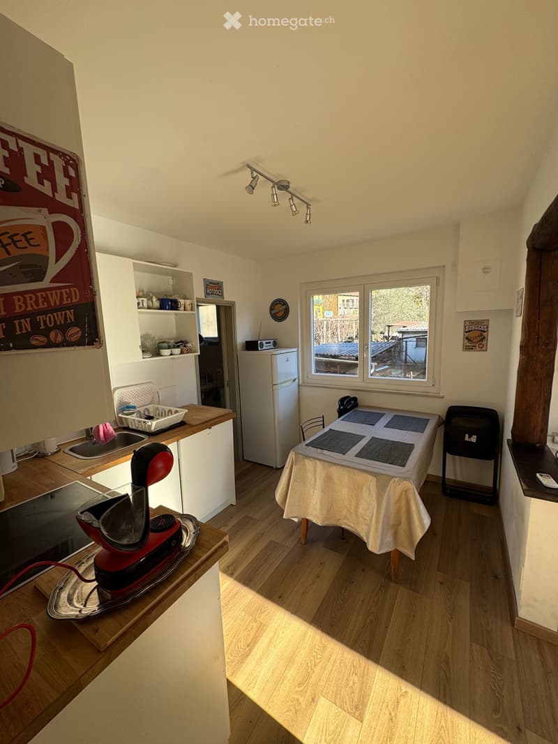 Compact kitchen with wooden flooring, white cabinets, a small refrigerator, a sink with a dish rack, a coffee maker on the counter, and a small dining table with chairs.