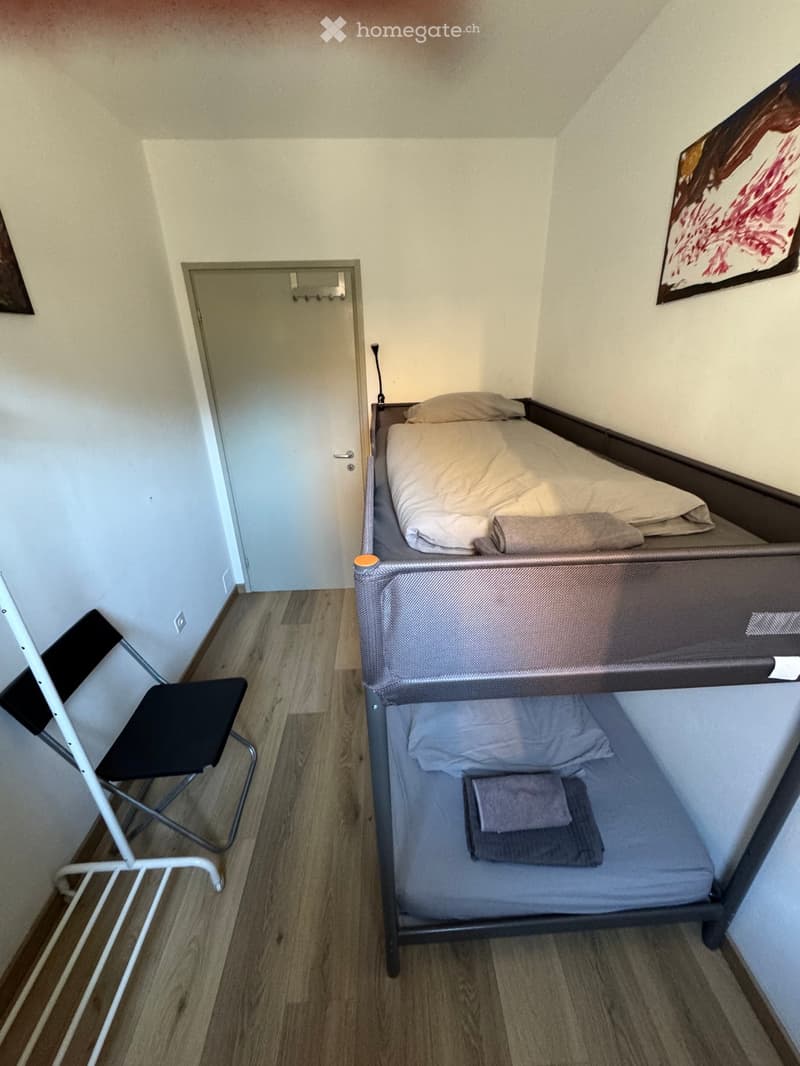 A small, furnished room with a bunk bed, a chair, and a door. The room has light-colored walls and wooden flooring.