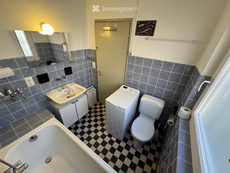 Bathroom with a sink, toilet, bathtub, washing machine, and a mirror. The walls are tiled in gray with a black and white checkered floor.