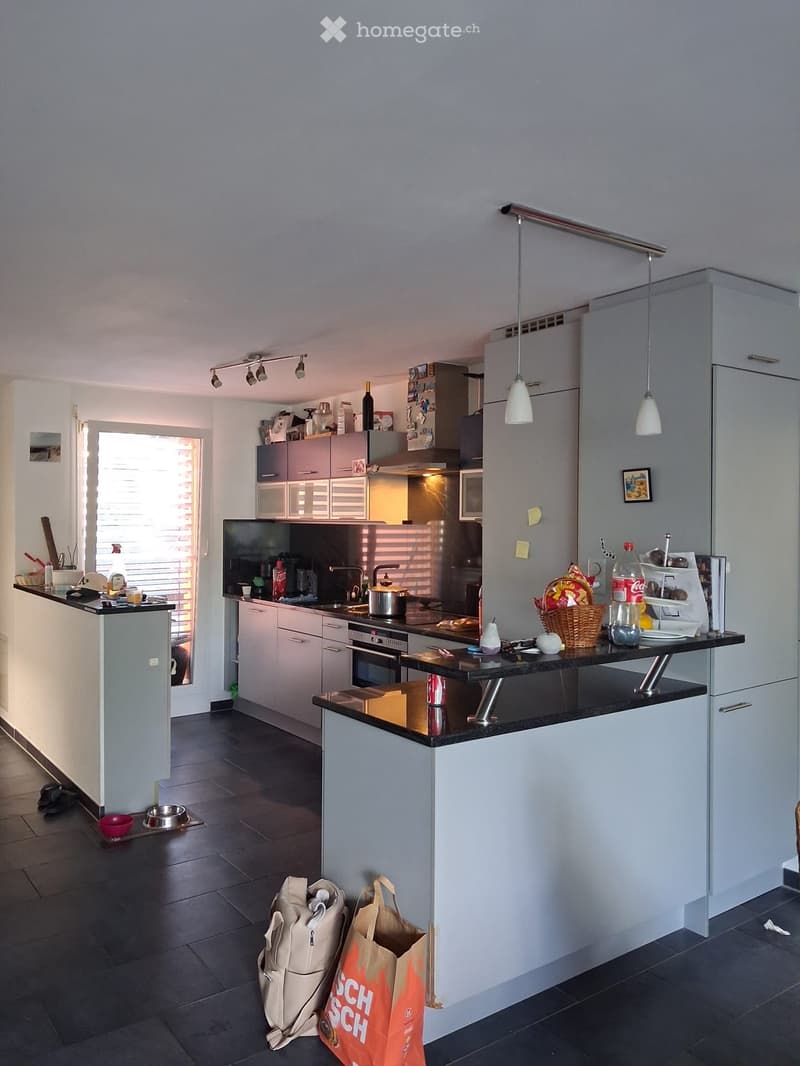 Modern kitchen with white cabinets, black countertop, stainless steel appliances, and pendant lights. There is a small island with a black countertop and a sink area with a faucet. The kitchen features a variety of items including a bottle, a basket, and