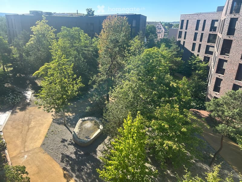 Aerial view of a landscaped garden area with trees, pathways, and a small water feature, surrounded by modern apartment buildings.