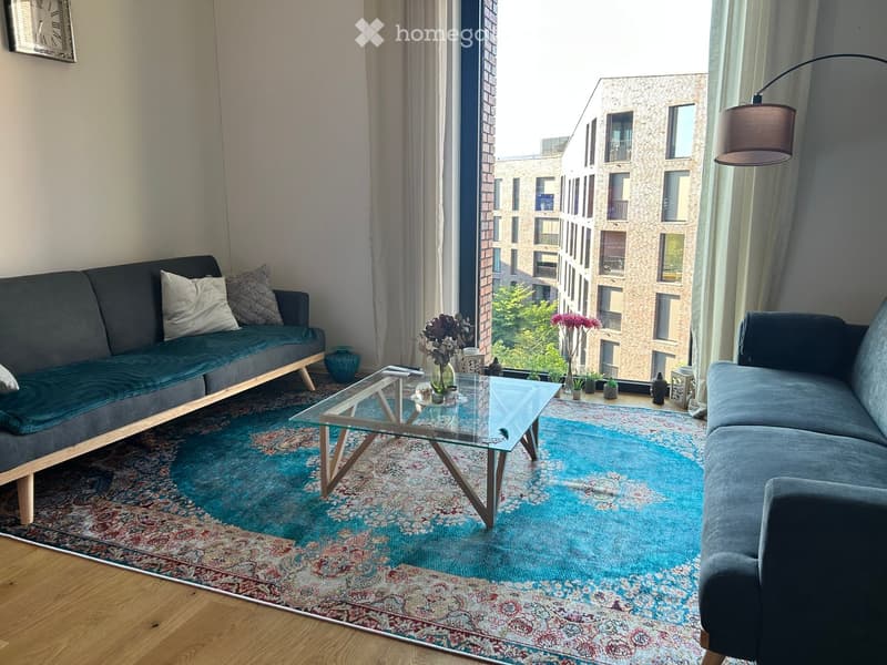 Modern living room with two sofas, a glass coffee table, a patterned rug, and a view of a cityscape through large windows.
