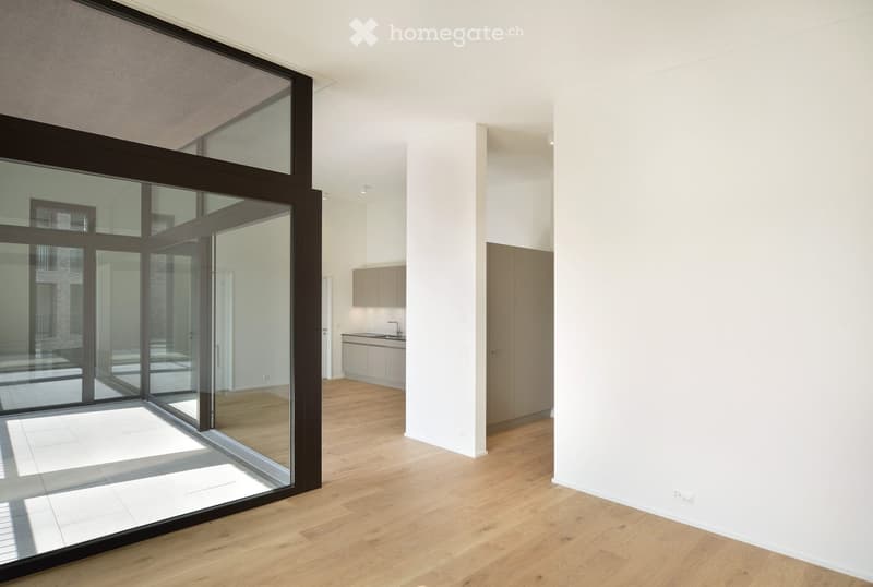 Bright, unfurnished room with large glass sliding doors leading to a tiled area, light wooden flooring, and a small kitchen area visible in the background.