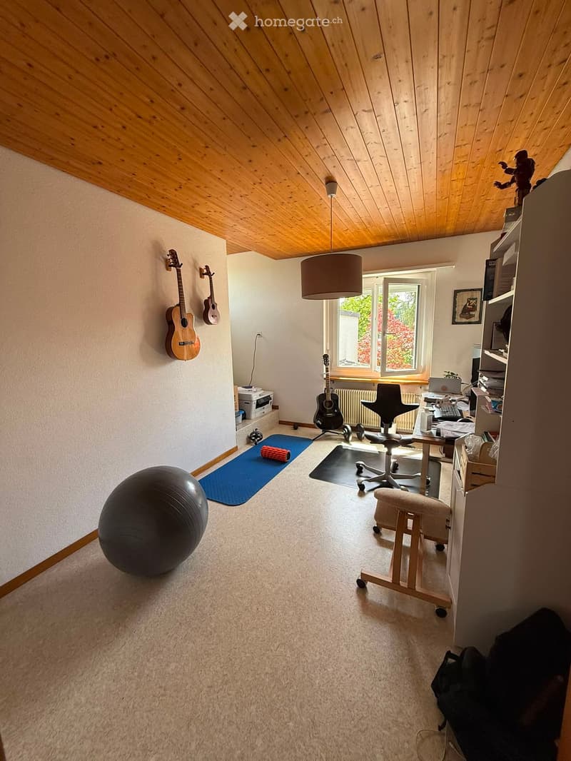 A multi-purpose room with a slanted wooden ceiling, large window, and various items including guitars, a ball, a chair, a table with a laptop, and a stool.