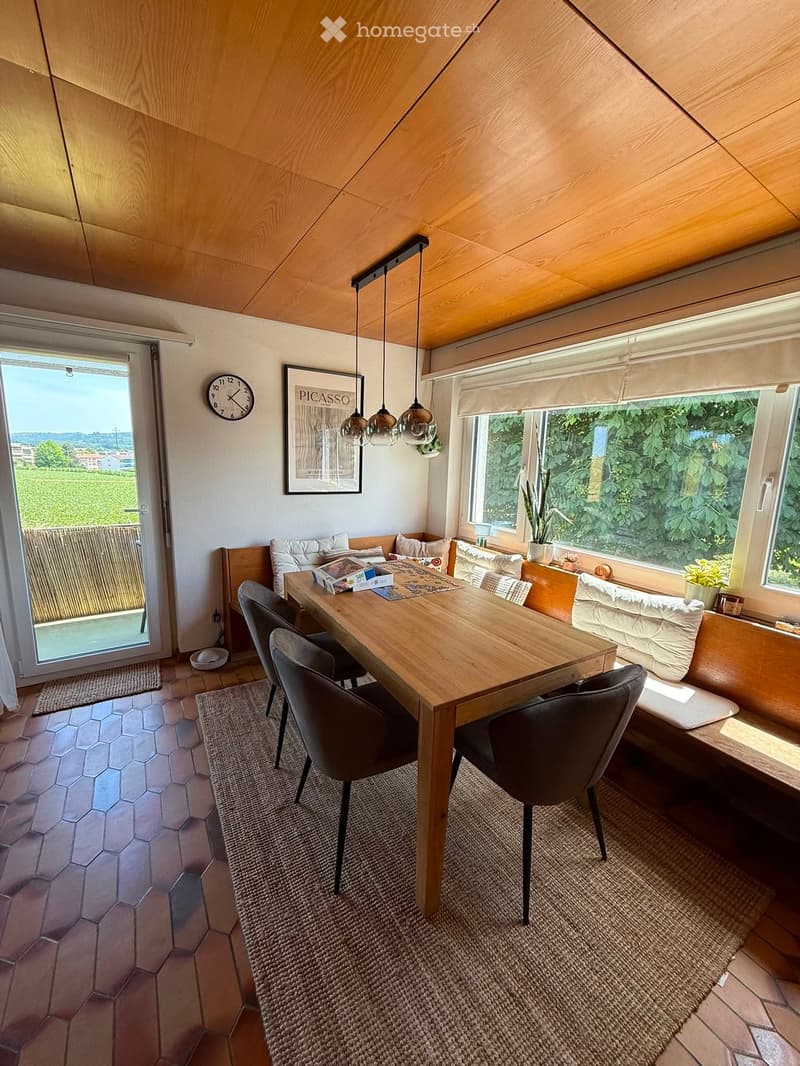 Bright, modern dining area with a wooden table, four chairs, a built-in sofa, pendant lights, a clock, and a picture frame on the wall. Large windows offer a scenic view of greenery.
