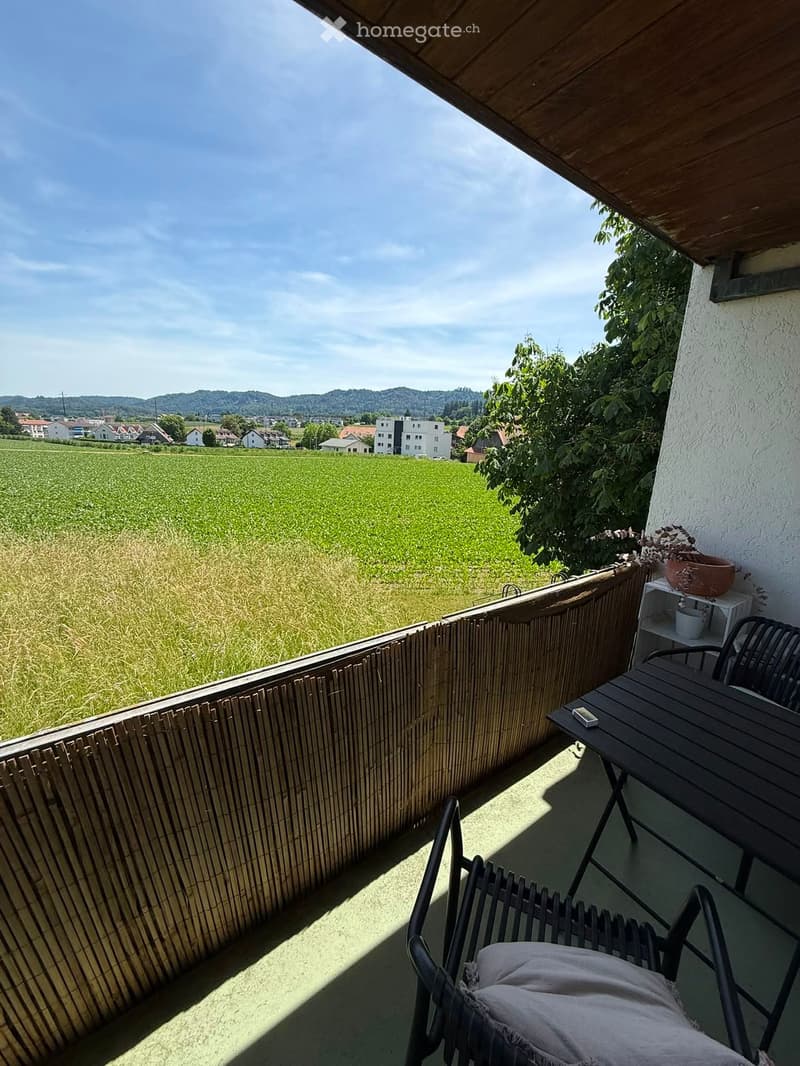 Balcony with a wooden railing, a small table, a chair with a cushion, and a cup on a shelf, overlooking a green field and distant houses.