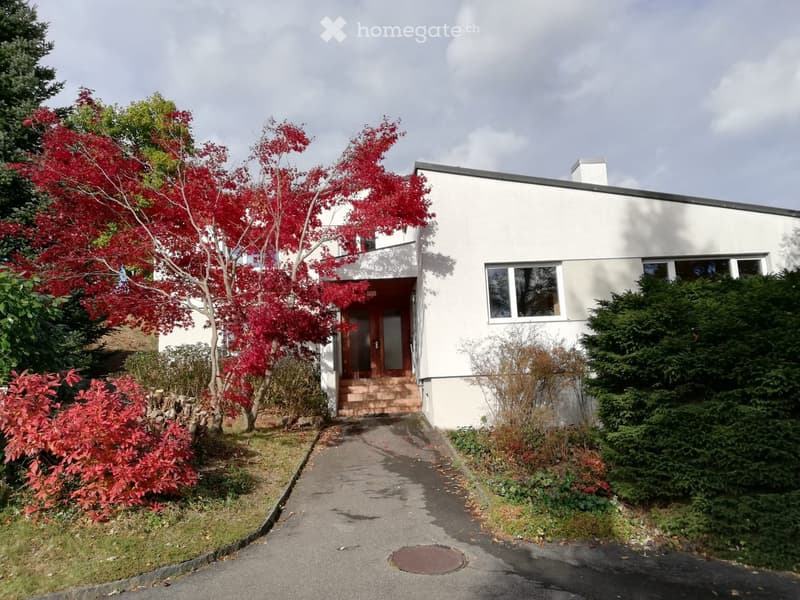 Modern two-story house with white exterior, large windows, and a small balcony. The entrance features a wooden door and steps leading up to it. The surrounding area includes a well-maintained garden with red-leaved trees and shrubs.