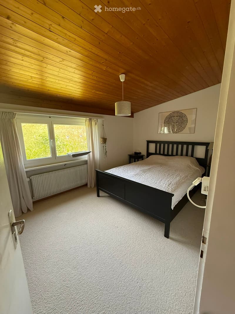 Bright bedroom with a wooden slanted ceiling, white walls, and large windows. Features a double bed with a dark wooden frame, a bedside table with a lamp, and a decorative picture frame on the wall.