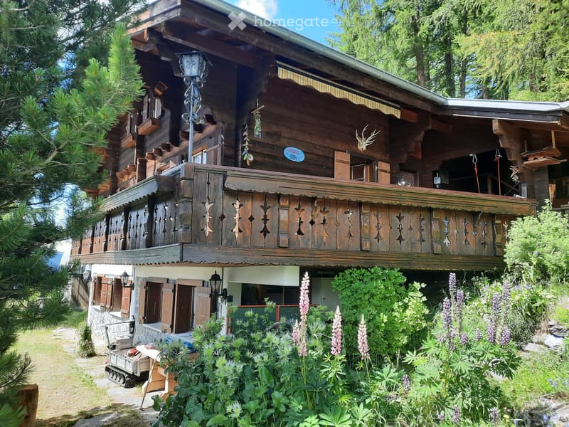 A two-story wooden house with a balcony, decorative wooden railings, and lush garden in front.