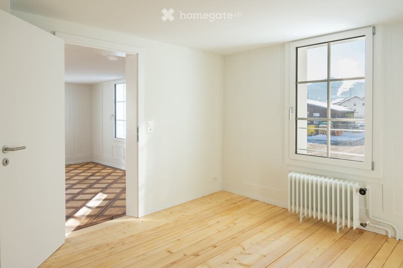 Empty room with wooden flooring, white walls, large window, radiator, and an open doorway leading to another room.