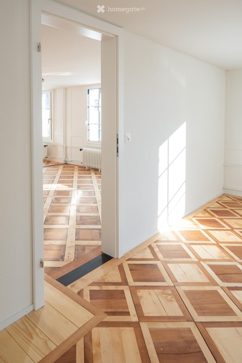 Empty hallway with wooden herringbone flooring, white walls, and a visible radiator in the background.