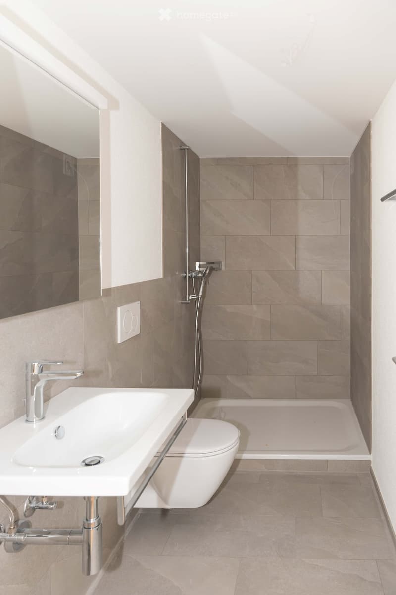 Modern bathroom with a white sink, chrome faucet, toilet, bathtub, and a large mirror. The walls are tiled in a light brown color, and the floor is covered with matching tiles.