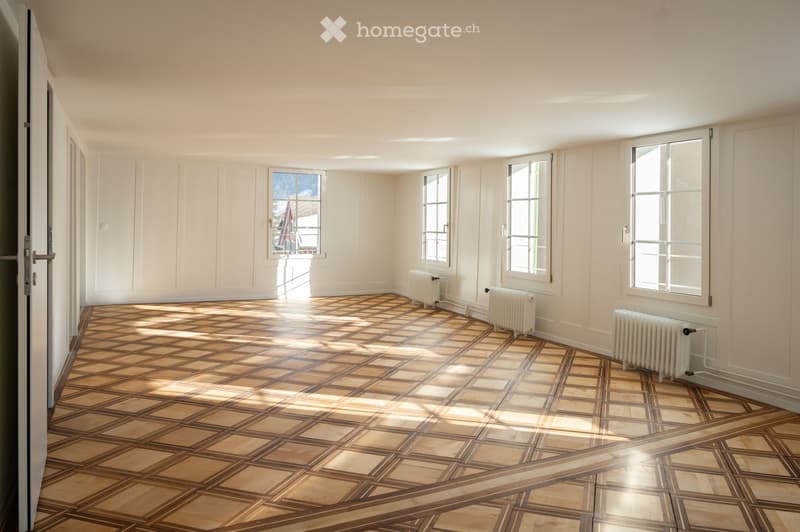 Empty room with light-colored walls, large windows, and a patterned wooden floor. Radiators are visible along the walls.