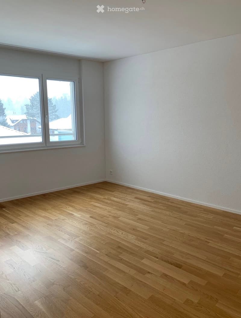 Empty room with light wooden flooring and white walls, featuring two large windows with a view of the outside.