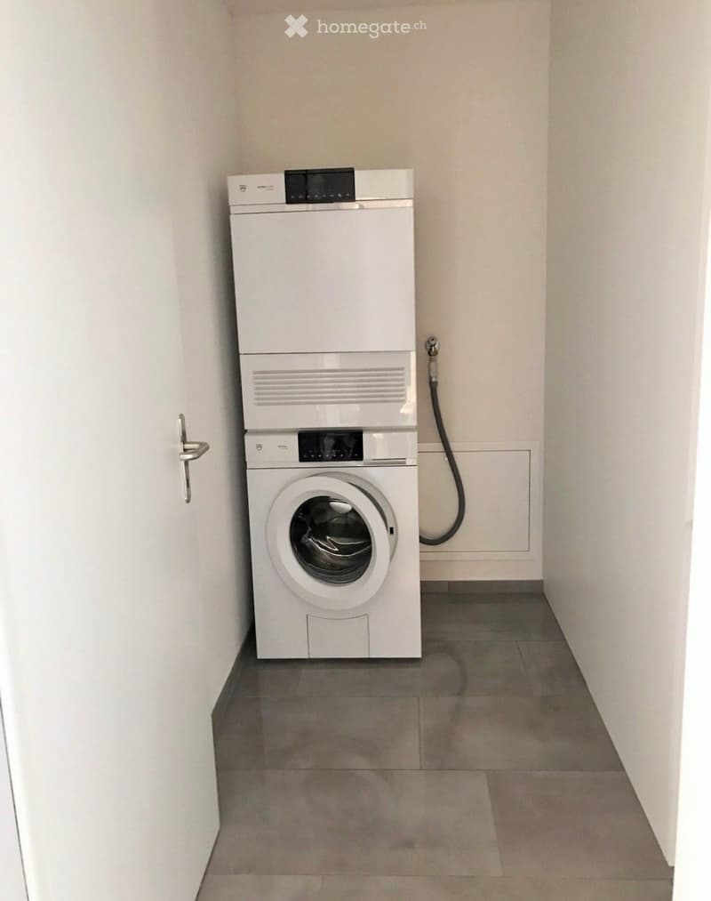 A small, clean room with a white washing machine and dryer stacked vertically, grey tiled floor, white walls, and a door with a silver handle.