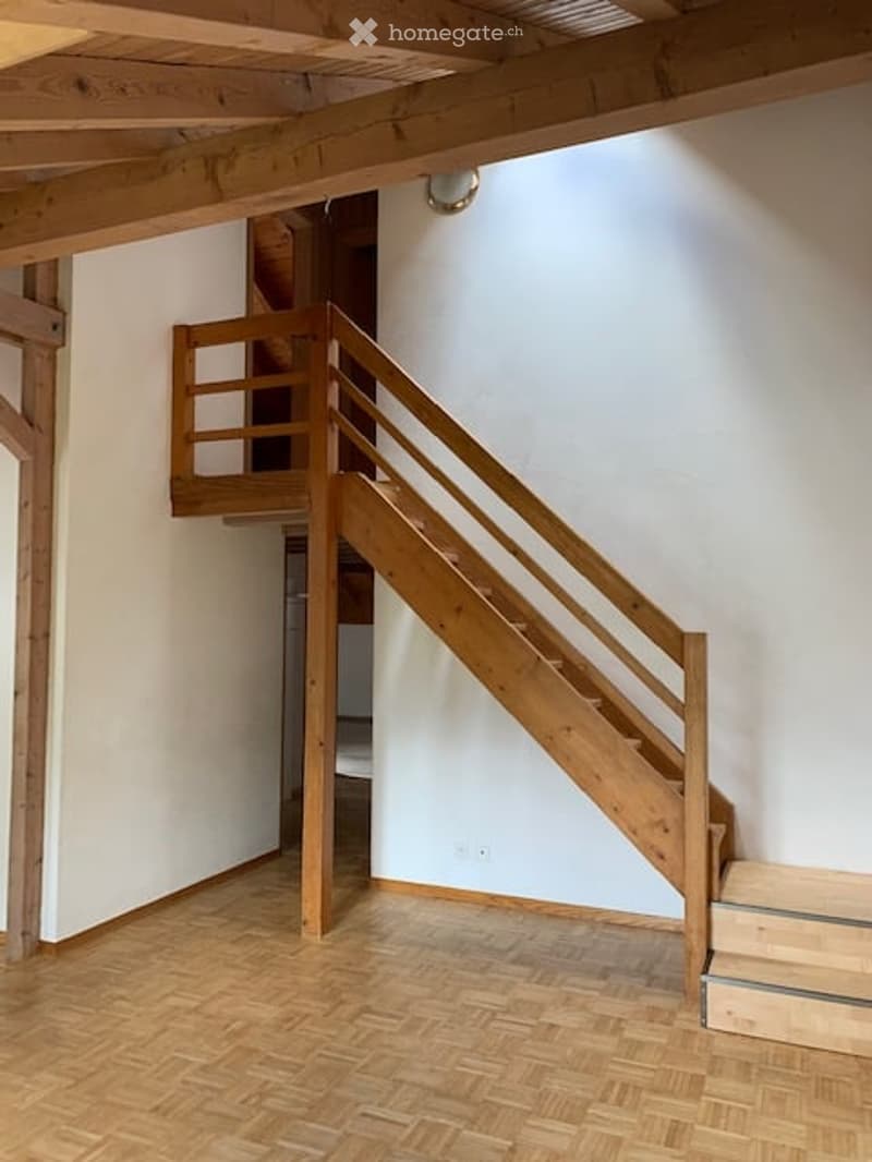 Wooden staircase with a simple wooden railing, leading to an upper level. The walls are painted white, and the floor is made of polished wooden planks.