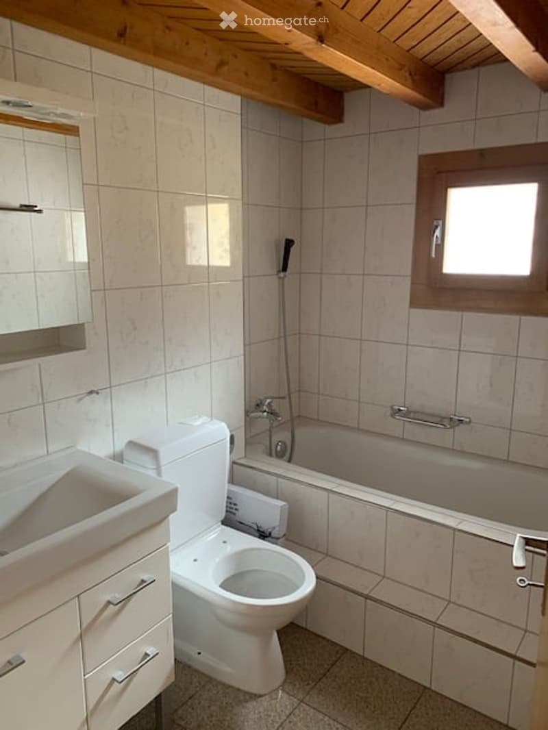 Bathroom with white tiles, a sink, toilet, bathtub, and a mirror above the sink. The room has wooden beams on the ceiling and a small window.