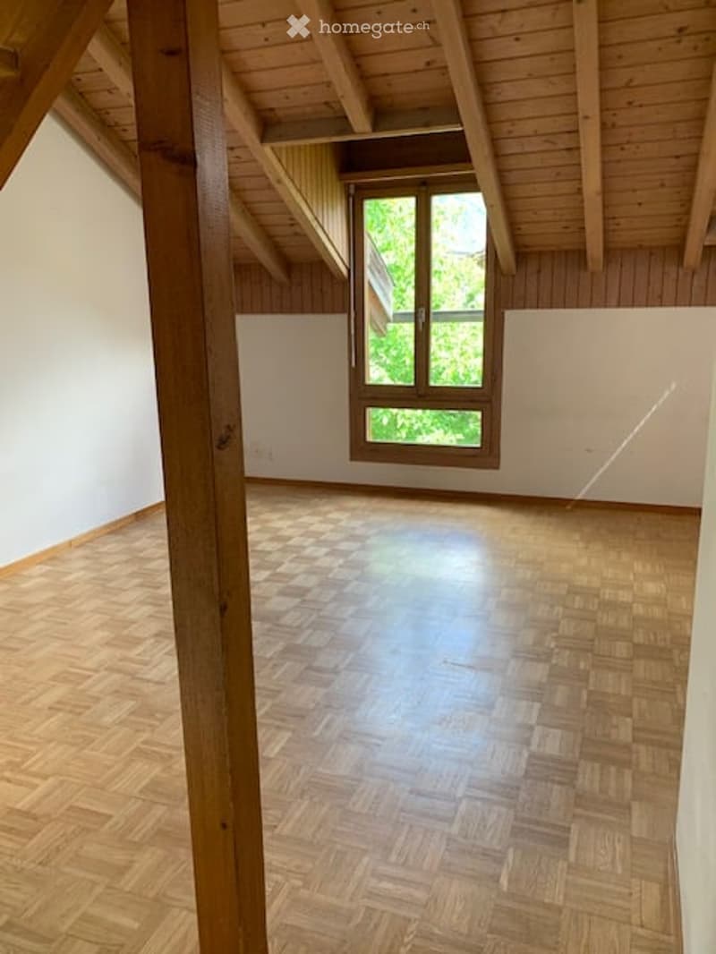 Empty room with wooden floor, white walls, and exposed wooden beams on the ceiling. A single window with wooden frame allows natural light to enter.