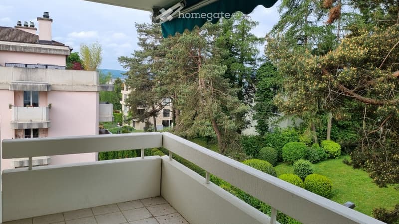 Balcony with white railings, tiled floor, overlooking a landscaped garden with trees and shrubs.