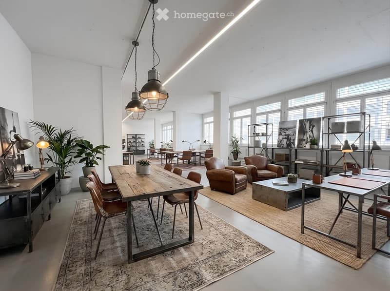 A spacious, well-lit room with a wooden dining table, leather armchairs, a bookshelf, and various decorative items. The room features large windows, pendant lights, and a cozy, industrial-style design.