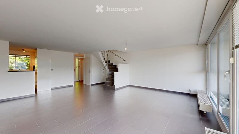 Empty room with white walls, dark tiled floor, and a staircase leading to another level. There are large windows on one side and a radiator under the windows.