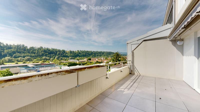 Spacious balcony with tiled flooring, white walls, metal railing, and scenic view of a green landscape and town below.
