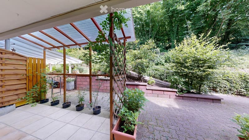 A covered outdoor patio area with a wooden frame and glass panels, surrounded by potted plants and lush greenery. The area features a tiled floor and is enclosed by a wooden fence and brick walls.