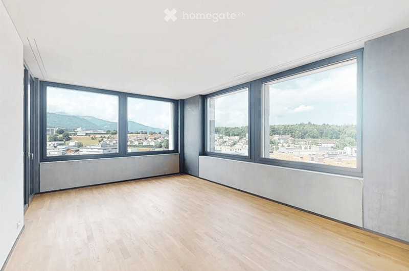 Empty room with wooden flooring, large windows offering a scenic view of a town and mountains, and grey walls.