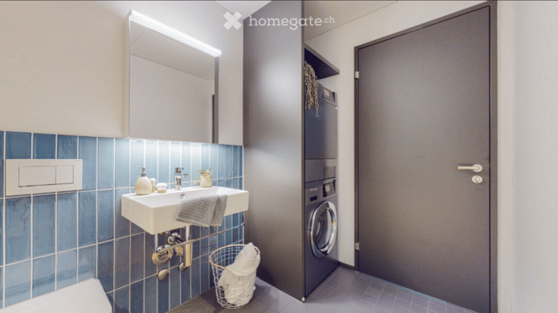 Modern bathroom with blue tiled walls, white sink, mirror, washing machine, and decorative items.