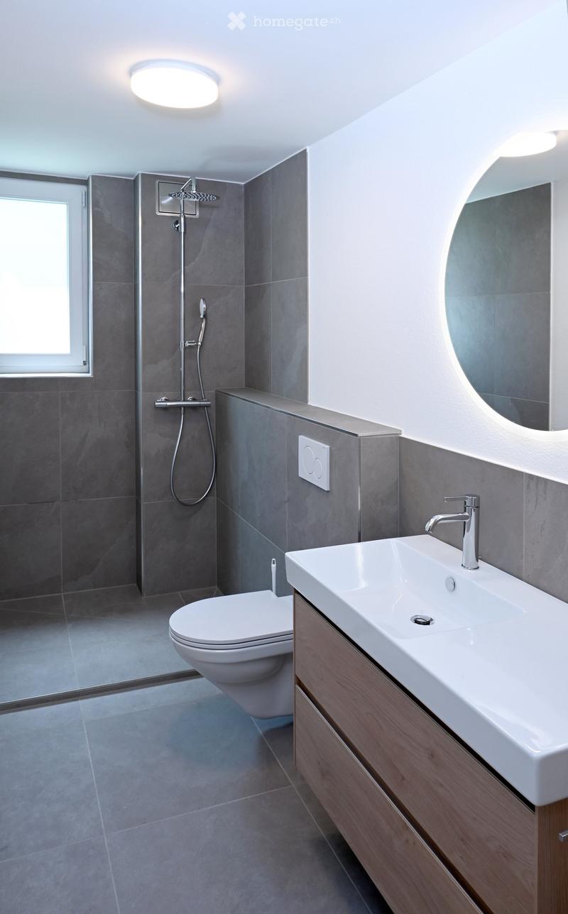 Modern bathroom with grey tiled walls, a white toilet, a white sink with a chrome faucet, a large round mirror, and a shower with a handheld sprayer.