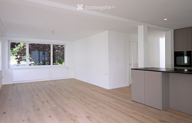 Empty room with light wooden flooring, white walls, large windows, and a grey kitchen island with an oven. No furniture or personal items present.