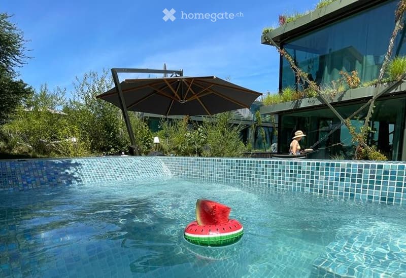 A modern swimming pool with a decorative watermelon float, surrounded by a sleek building with large glass windows and a green roof. A large umbrella provides shade, and the area is set in a lush, green environment.