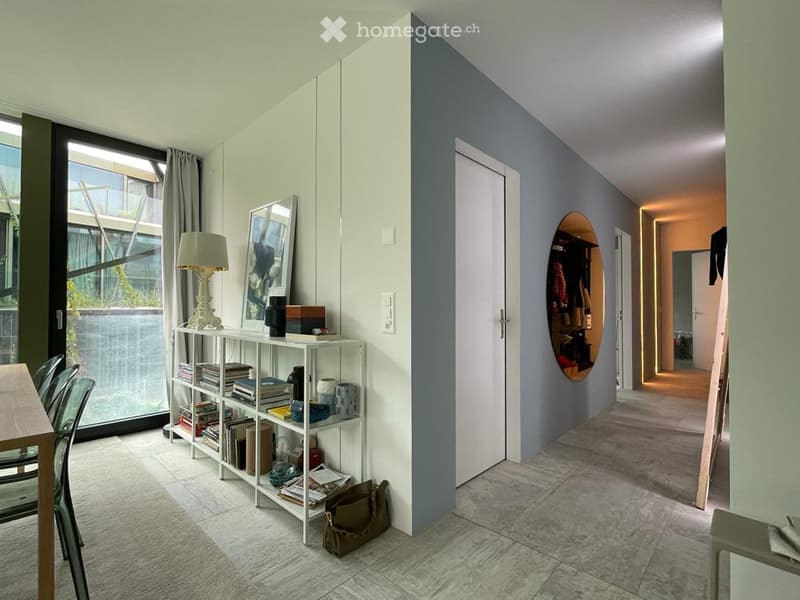 Modern hallway with a white shelf, a small table, chairs, a lamp, and a mirror. The hallway leads to a room with a visible balcony.
