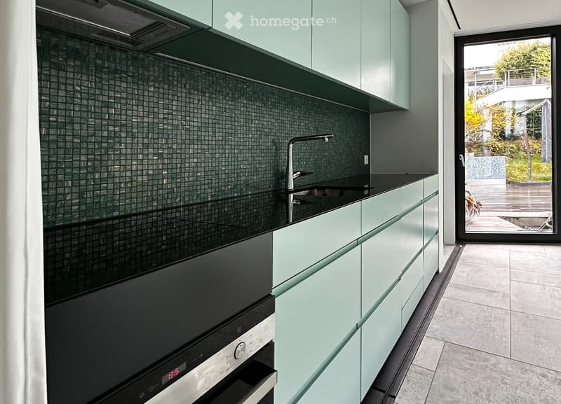 Modern kitchen with light green cabinets, black countertop, and a built-in oven. Features a sleek faucet and a tiled backsplash.
