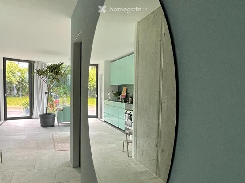 Modern open-plan living and kitchen area with light green cabinets, a sofa, a small stool, and a large potted plant near the sliding glass doors.