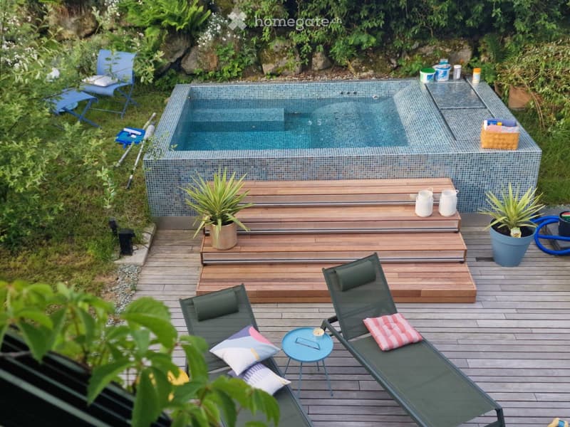A small, modern swimming pool surrounded by wooden decking, with lounge chairs, a small table, and potted plants. The area is enclosed by greenery and features a wooden staircase leading to the pool.