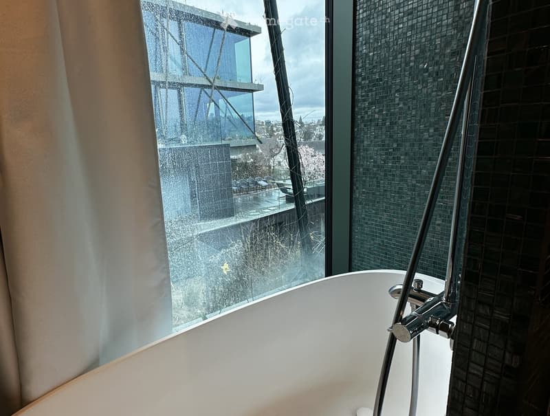Modern bathroom with a freestanding bathtub, sleek faucet, and large window offering a view of a modern building and outdoor seating area.