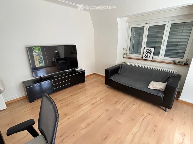 Modern living room with light wooden flooring, a black leather sofa, a TV stand with a large flat-screen TV, a black chair, and a mirror on the wall.