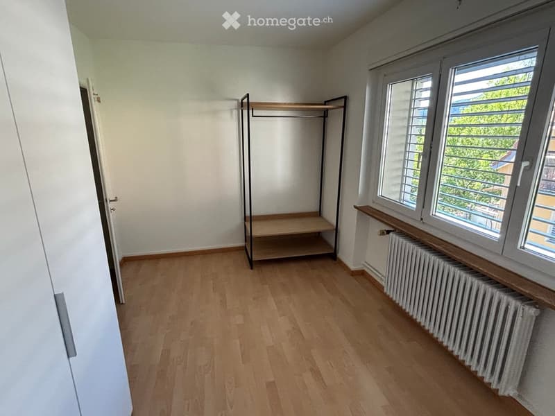Empty room with wooden flooring, white walls, large windows with blinds, and a simple clothing rack with shelves.