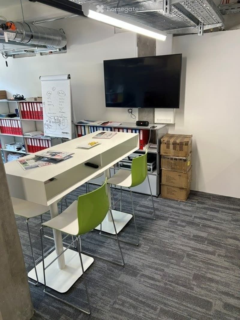Modern office space with a white table, green chairs, a large screen on the wall, shelves with books and files, and stacked storage boxes.