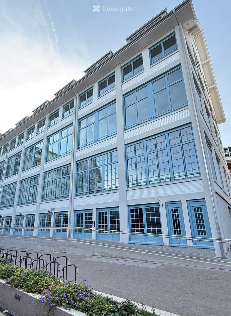Modern multi-story building with large windows and blue-framed doors, featuring a raised ground floor and a metal railing along the sidewalk.
